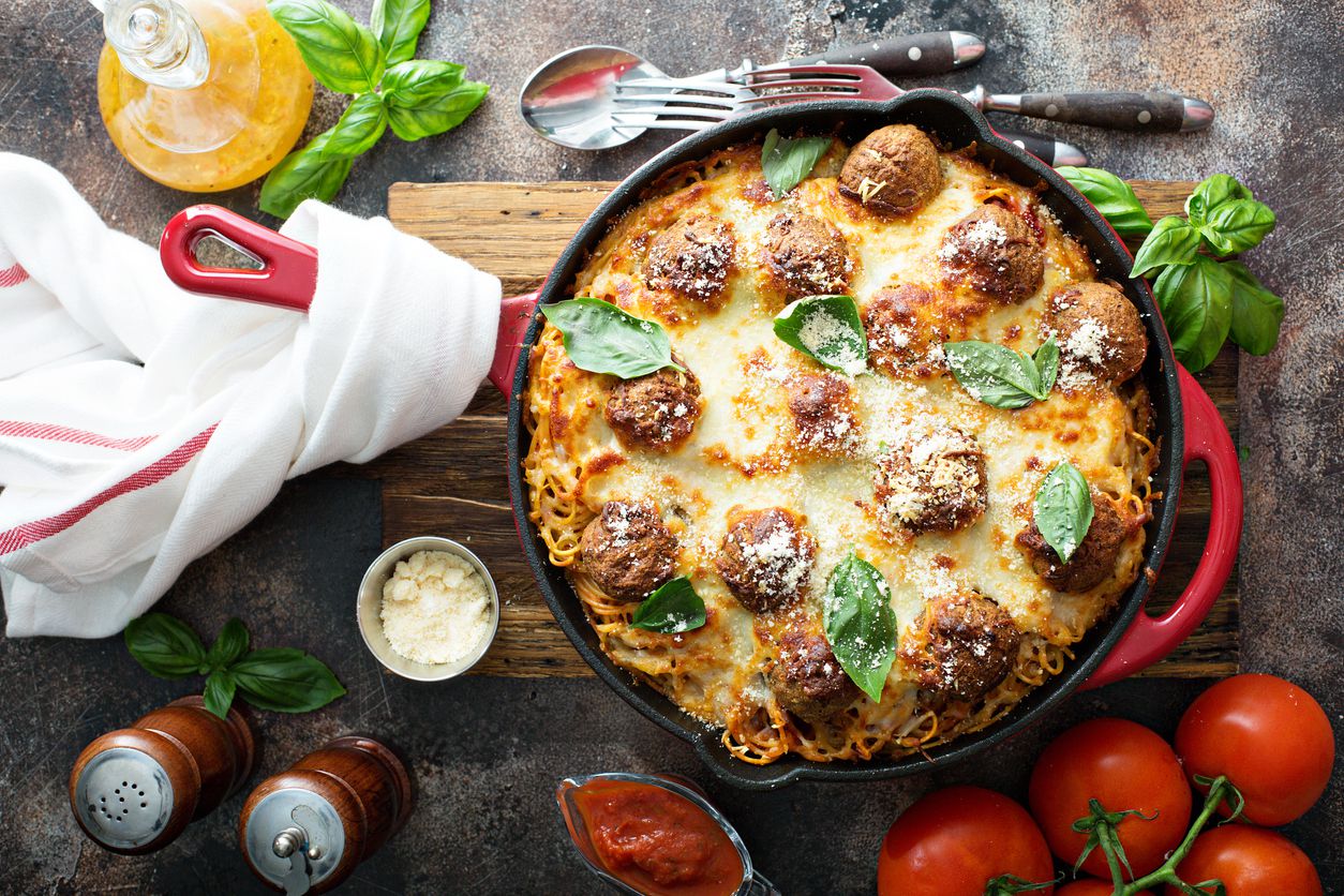 Baked meatballs in cast iron pan on a table with salt and pepper shaker, tomatoes on the vine, parmesan cheese, basil and cutlery