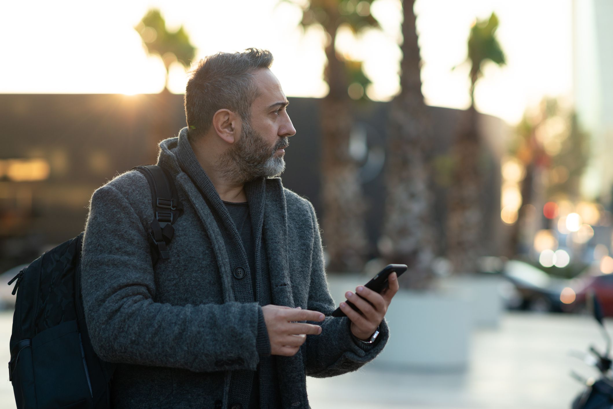 Worried man looking thoughtfully away. He is using a smart phone outside corporate building.