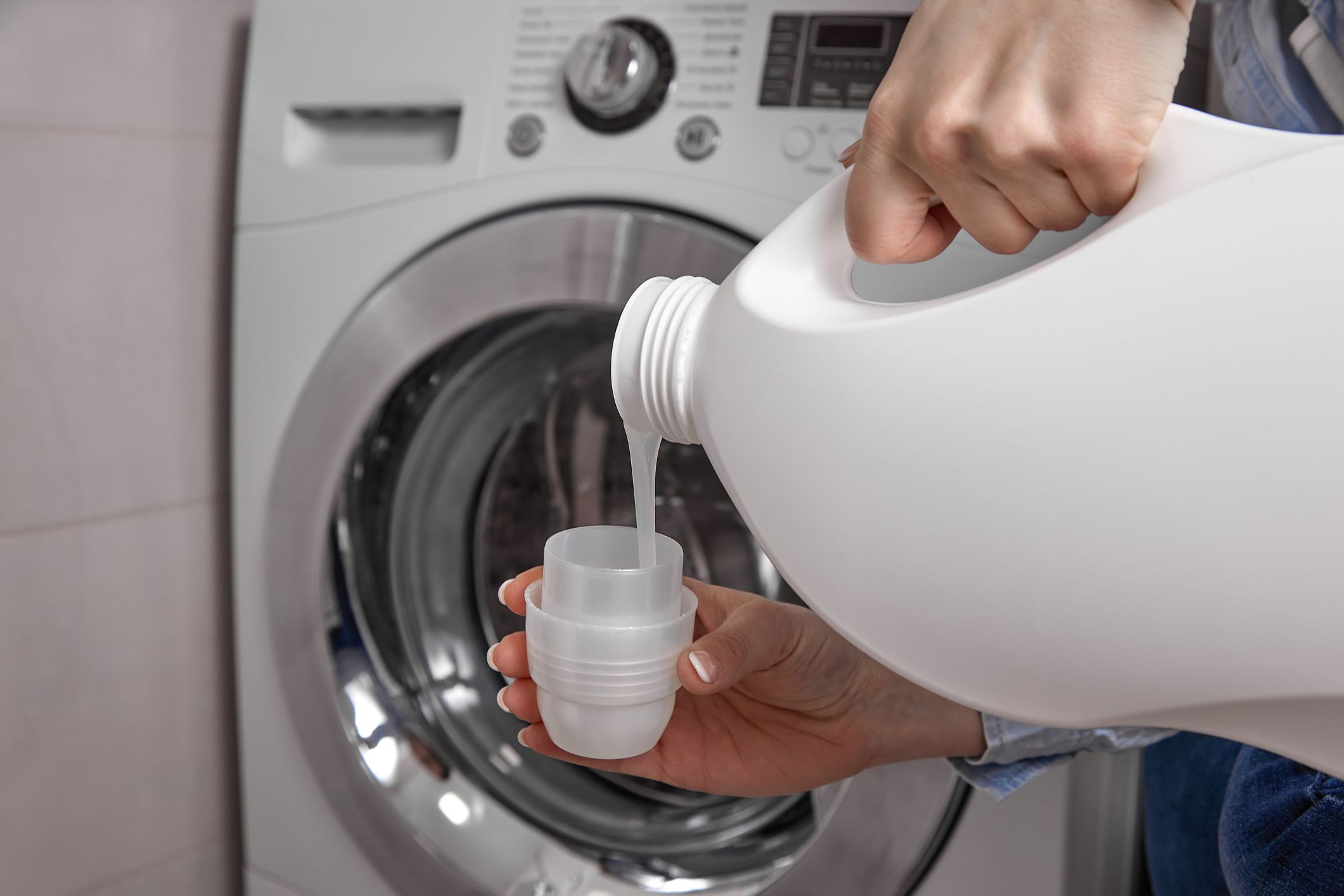 In bathroom where washing machine is located, girl pours the laundry conditioner into lid to give clean laundry pleasant smell and softness. From white plastic bottle, rinse is poured into the cap