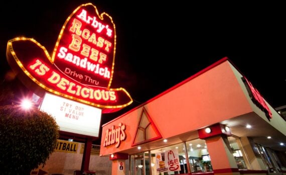 A brightly lit Arby's restaurant at night, featuring a large neon cowboy hat sign that reads "Arby's Roast Beef Sandwich is Delicious" above the drive-thru. The building has red trim and large windows.