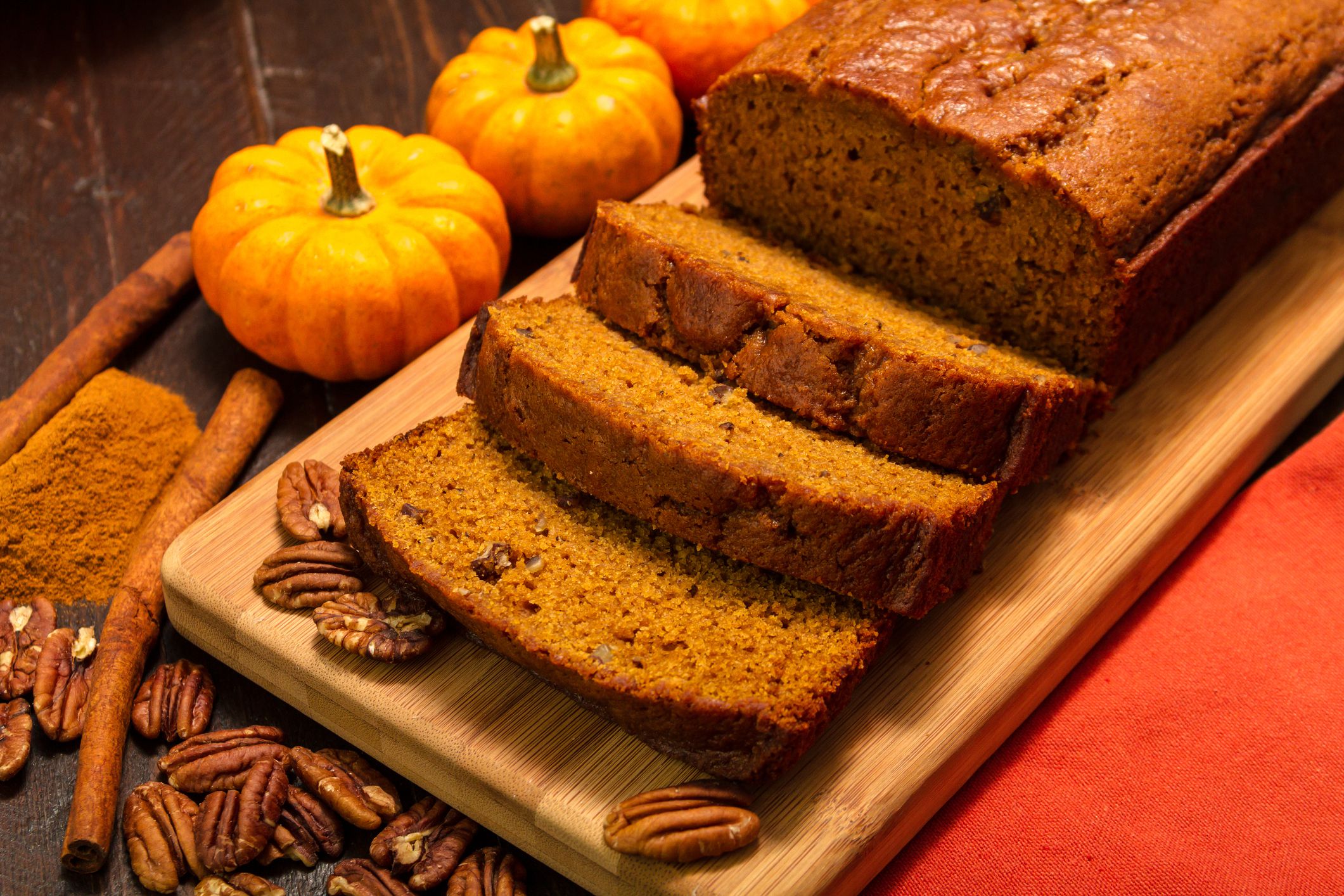 Festive setting of sliced pumpkin loaf sitting on wooden cutting board with orange cloth napkin, cinnamon sticks and pecan nuts