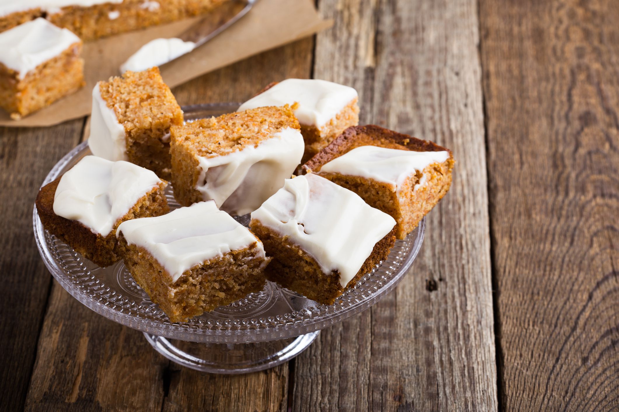 Homemade spiced pumpkin cake with cream cheese frosting sliced into squares, served on glass stand on rustic wooden table