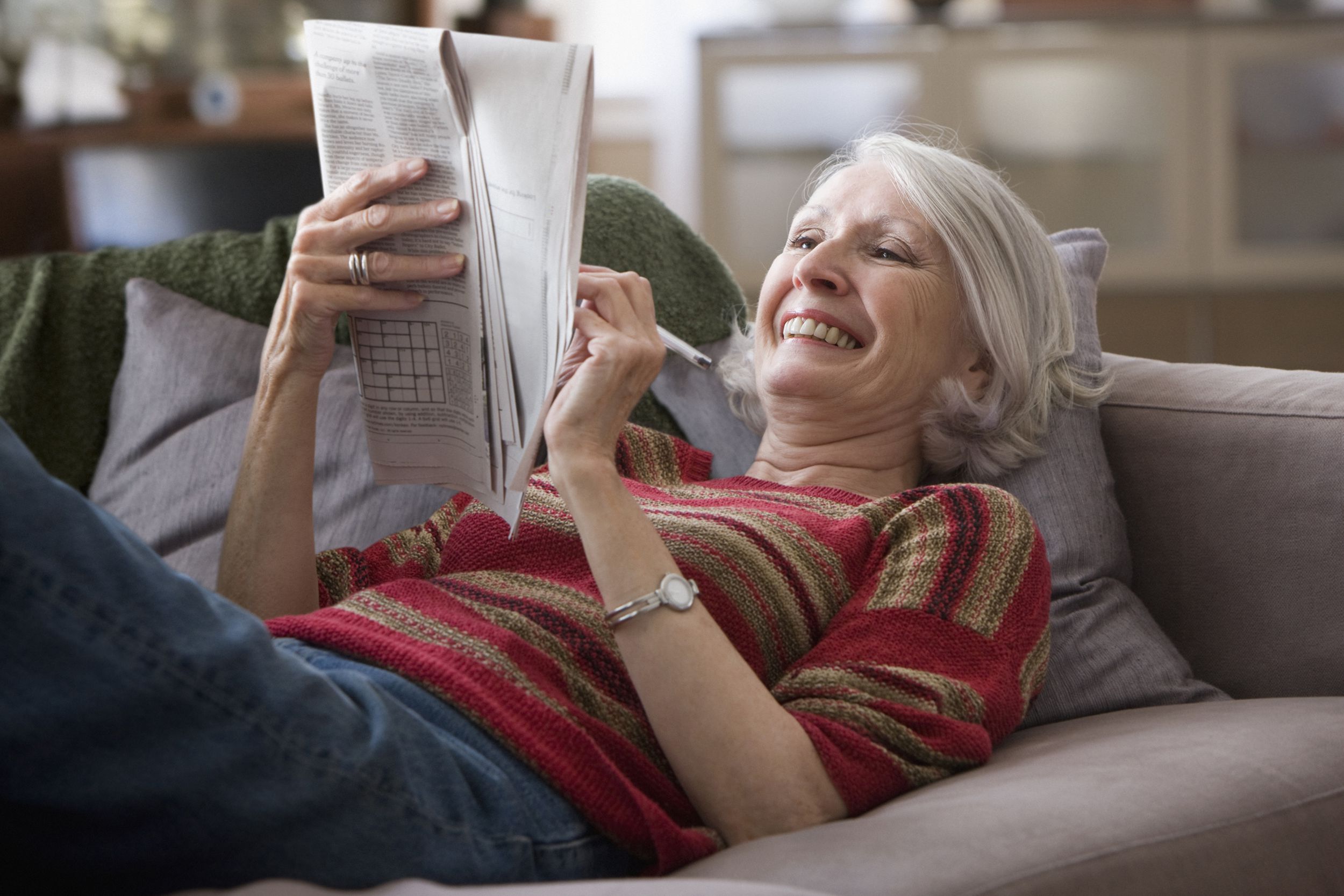 senior woman laying down on couch in living room looking over newspaper