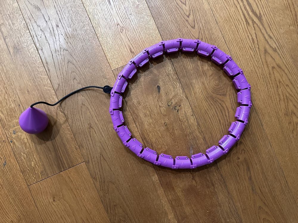 A purple weighted hula hoop with adjustable segments and a black cord attached to a weighted ball, lying on a wooden floor.