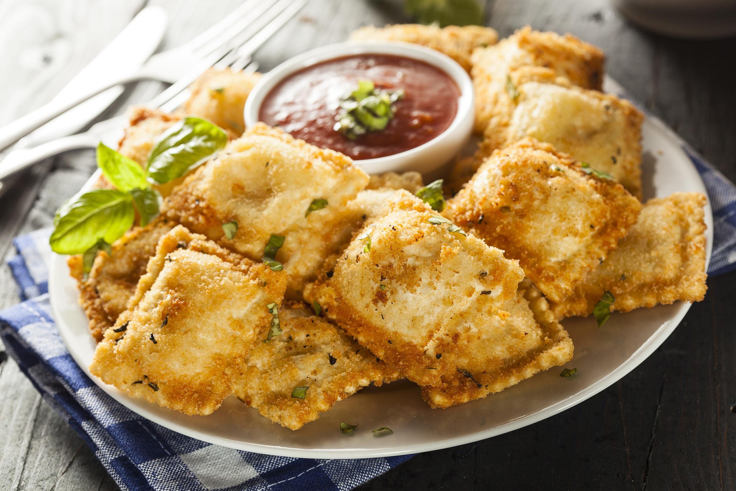 A pile of fried raviolis on a white plate with a small round bowl of marinara sauce in the middle on a blue and white checkered napkin on a wooden table with utensils in the background