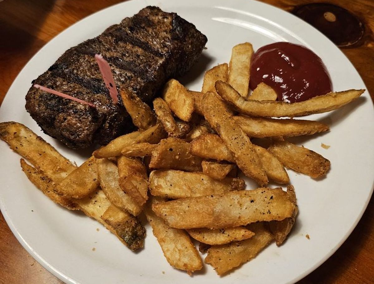 A white plate with grilled steak—like the kind served at cheap steak restaurants—seasoned French fries, and a serving of ketchup sits on a wooden table. Two pink toothpicks are inserted into the steak.
