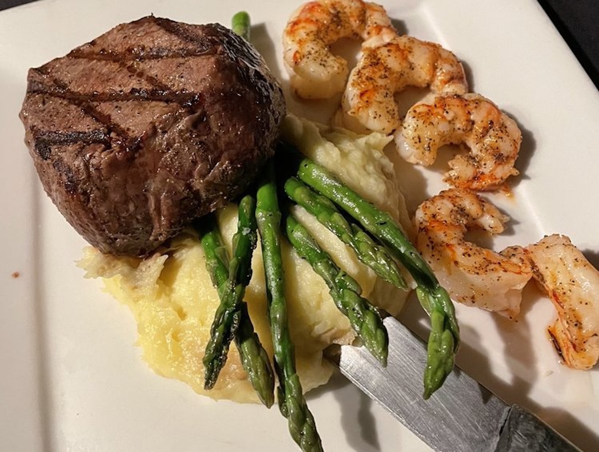 A plate with grilled steak, mashed potatoes topped with asparagus spears, and seasoned shrimp—reminiscent of dishes at cheap steak restaurants. A knife rests beside the potatoes on a neatly arranged white square dish.