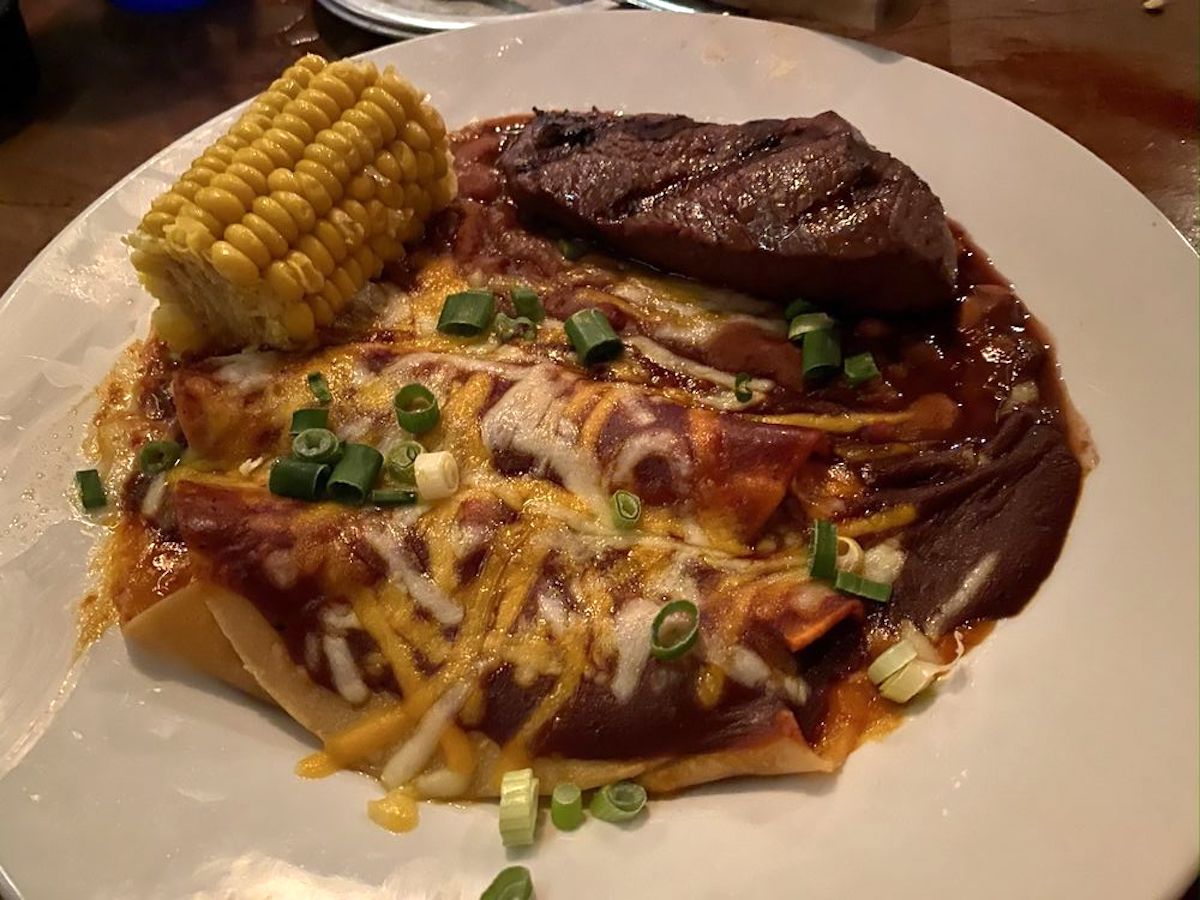 A plate with enchiladas topped with melted cheese and chopped green onions, a grilled steak like those at cheap steak restaurants, and a piece of corn on the cob.