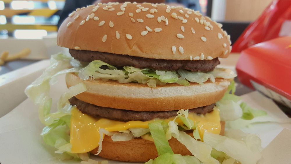 A close-up of a sesame seed hamburger bun with two beef patties, shredded lettuce, slices of cheese, and sauce, resembling a classic fast food burger.