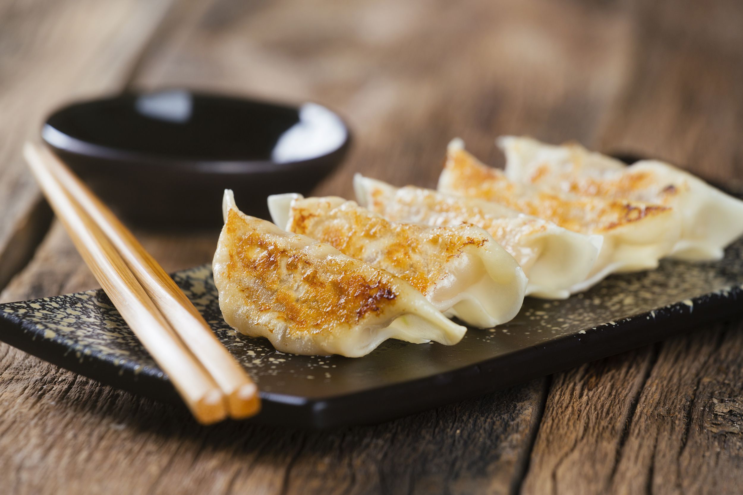 Closeup of five dumplings with chopsticks on a black rectangle plate with a small black bowl of dipping sauce on a wooden table with a blurred background