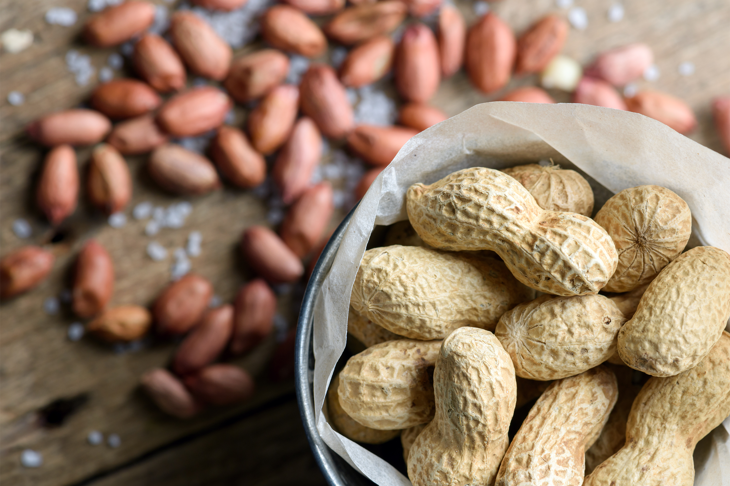 Closeup of shelled peanuts in a wax paper lined tin bucket on the right side, surrounded by a blurred background of shelled peanuts on a rustic wooden table