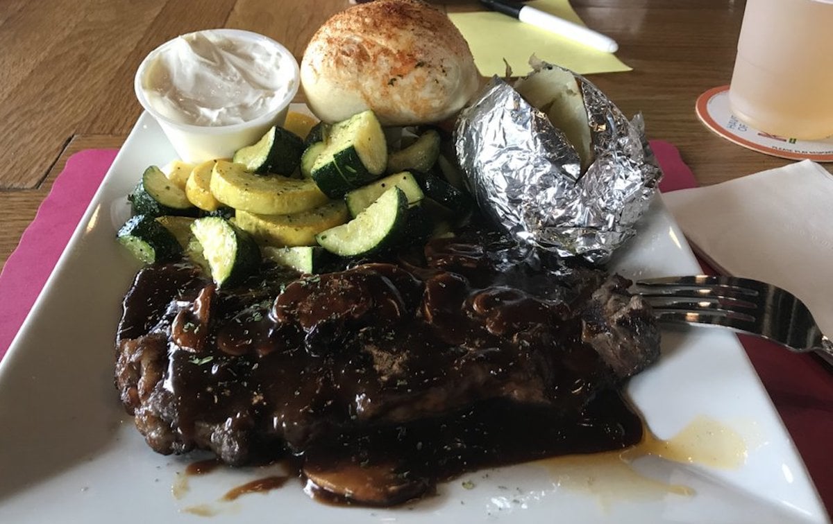 A plate with grilled steak topped with mushroom gravy, sliced sautéed zucchini and yellow squash, a baked potato in foil, a bread roll with butter on the side—just like the hearty meals served at cheap steak restaurants.