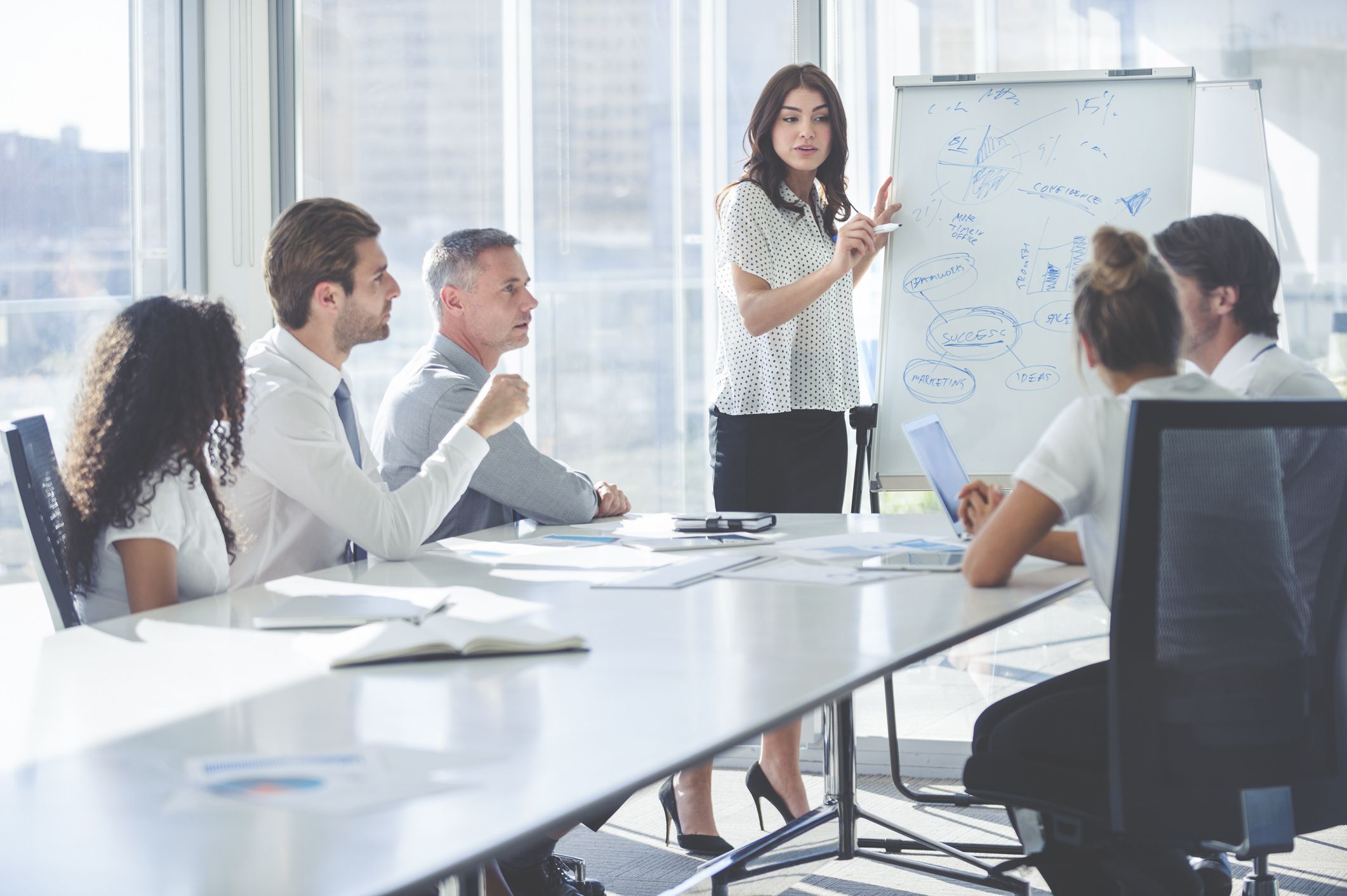 Female market research analyst giving a presentation while standing with a whiteboard to her team while in a conference room with sunlight coming through the windows in a city