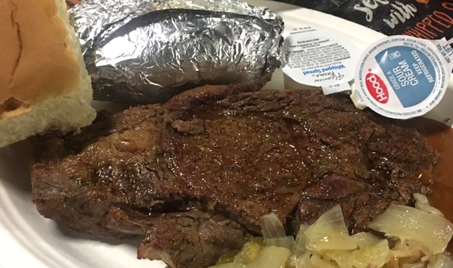 A classic plate from cheap steak restaurants: grilled steak, baked potato wrapped in foil, a fluffy bread roll, savory cooked onions, and two containers of sour cream.