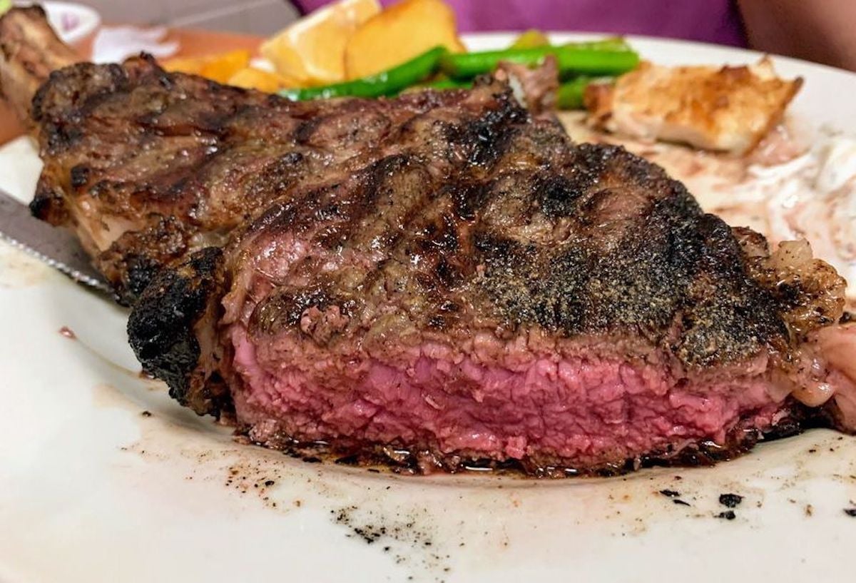 A thick, juicy steak cooked medium rare, sliced to show a pink center, sits on a white plate with grill marks—just what you'd expect from the best cheap steak restaurants. Some asparagus and roasted potatoes are visible in the background. Ray's Cafe in Hawaii