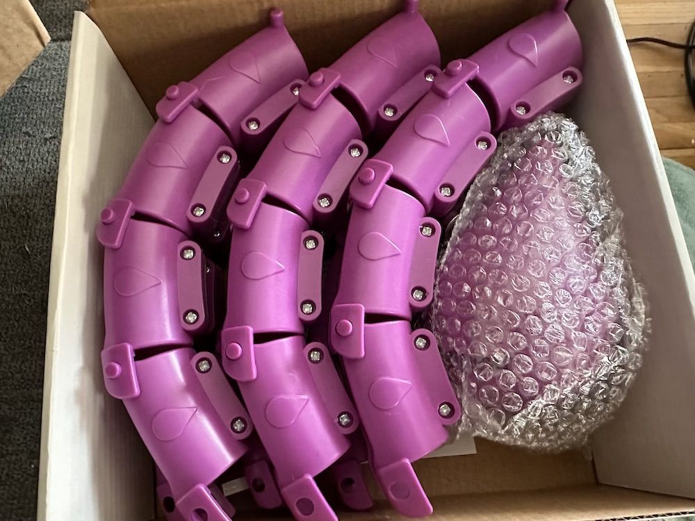 Several curved, purple plastic pieces with screws are neatly arranged in a box alongside a bubble-wrapped item. The purple pieces have a teardrop-shaped embossing on each segment.