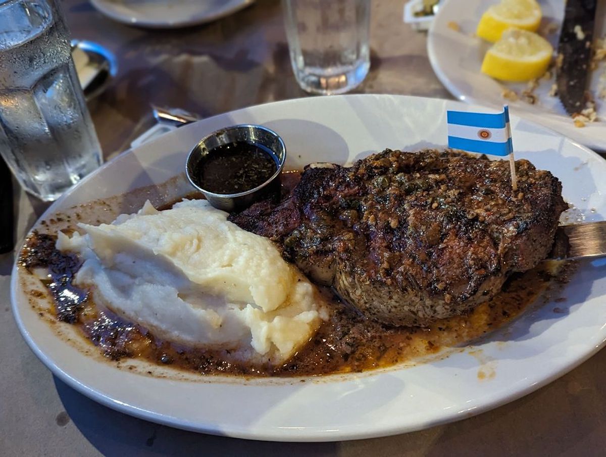 A plate with mashed potatoes, a grilled steak topped with herbs and a small Argentinian flag—typical fare at cheap steak restaurants—a cup of dark sauce, water, and lemon wedges in the background.