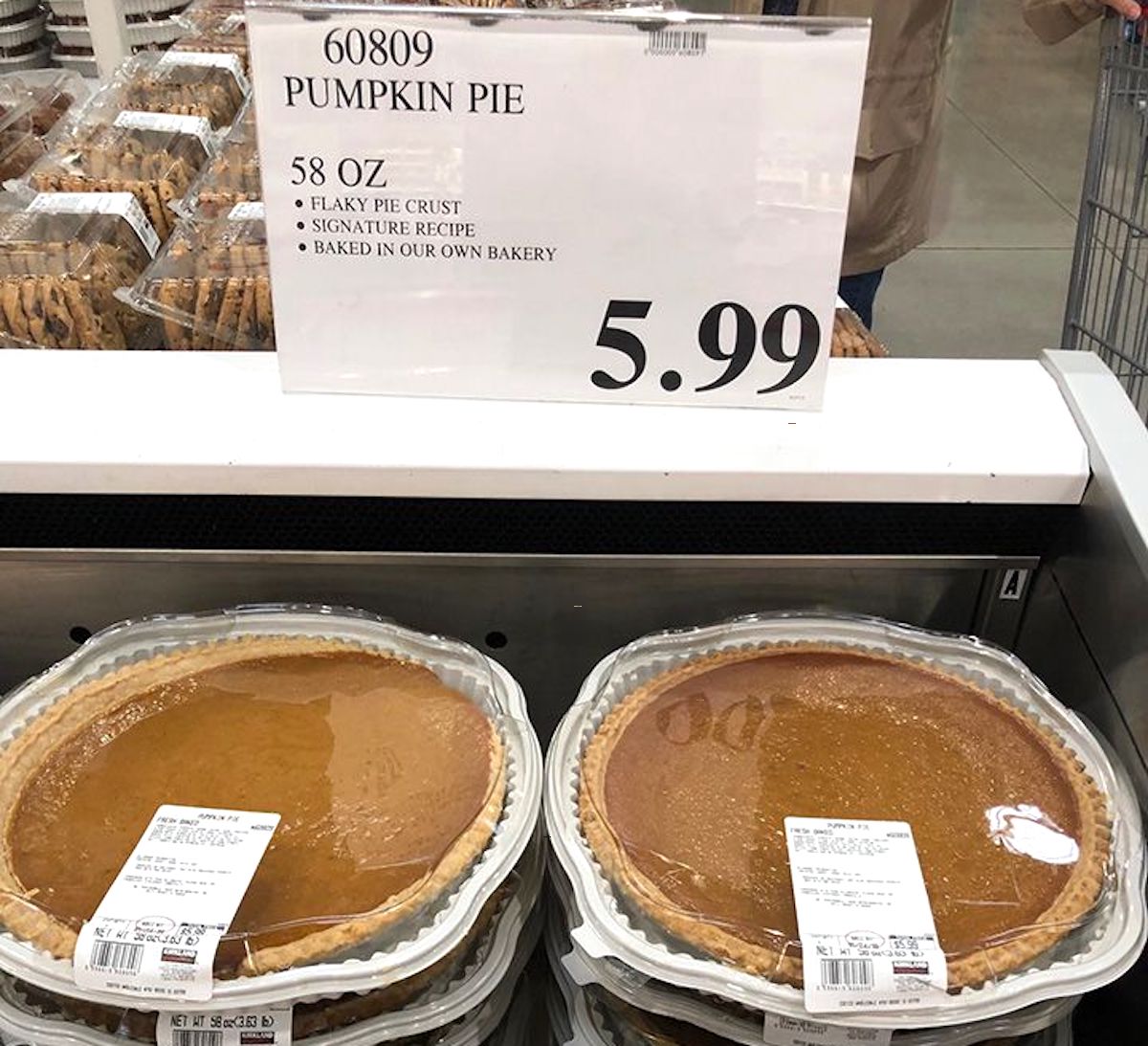 Two large pumpkin pies in clear plastic containers sit on a store shelf under a sign reading “Pumpkin Pie, 58 oz, $5.99,” making them perfect Costco Thanksgiving sides. Other bakery items can be seen in the background.