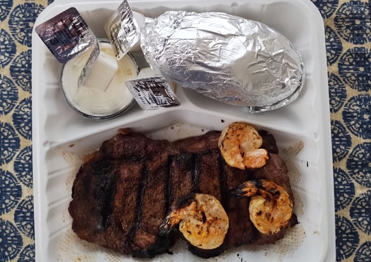 A takeout container from one of the best cheap steak restaurants holds a grilled steak, three grilled shrimp, a foil-wrapped baked potato, and containers of sour cream and butter on a patterned blue and cream tablecloth.