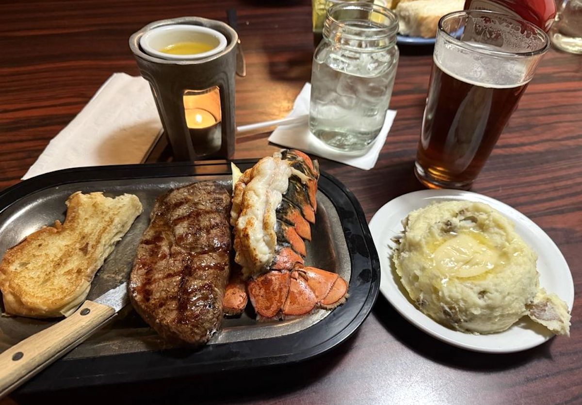 A surf and turf meal with grilled steak, lobster tail, and garlic bread on a metal plate, served with mashed potatoes, a glass of beer, and water on a wooden table—similar to what you’d find at cheap steak restaurants. A candle burns in a holder nearby.