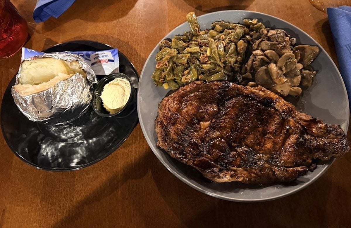 A grilled steak with sautéed mushrooms and green beans sits on a gray plate, served alongside a baked potato wrapped in foil, butter, and sour cream—reminiscent of meals at cheap steak restaurants. A blue napkin rests on the wooden table.