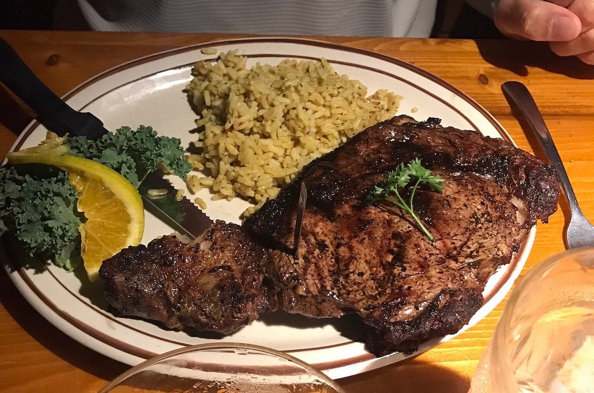 A plate with a large grilled steak garnished with parsley, seasoned rice, kale, and an orange slice evokes the hearty meals found at cheap steak restaurants, set invitingly on a wooden table with utensils.