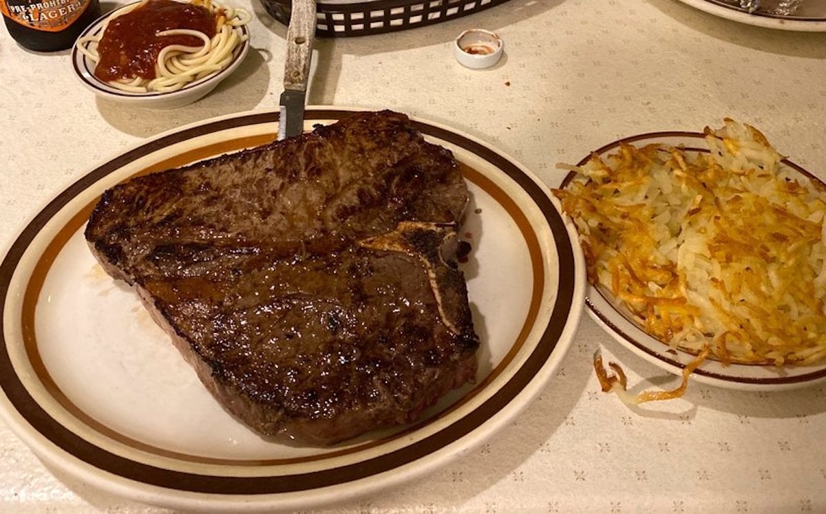 A large grilled T-bone steak on a plate with a knife in it, next to crispy hash browns—this hearty meal is just what you’d expect at the best cheap steak restaurants. In the background, there’s a small plate of spaghetti with tomato sauce.