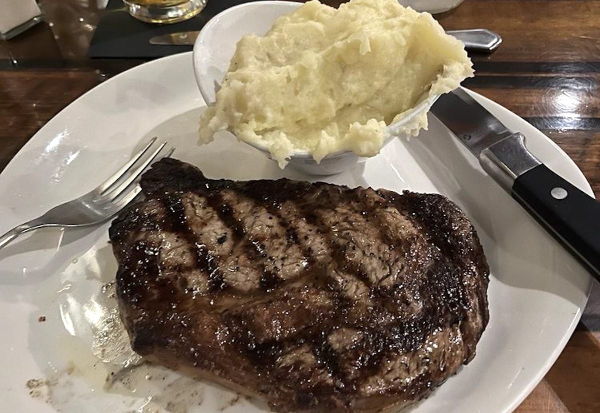 A grilled steak with sear marks sits on a white plate next to creamy mashed potatoes. Perfect for fans of cheap steak restaurants, this hearty meal is served on a wooden table with a fork and steak knife ready to dig in.