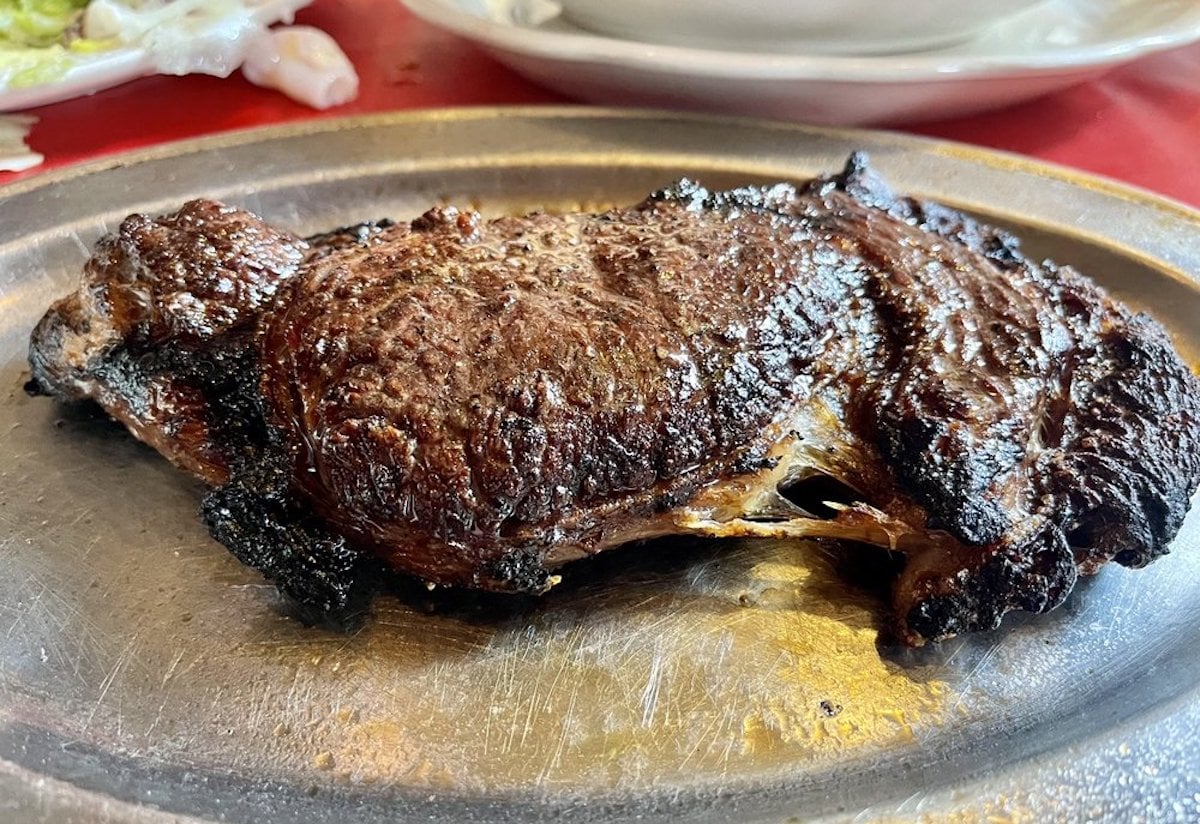 A grilled steak with a charred exterior sits on a metal plate, capturing the mouthwatering appeal often found at cheap steak restaurants, with parts of a salad and a white plate visible in the background.