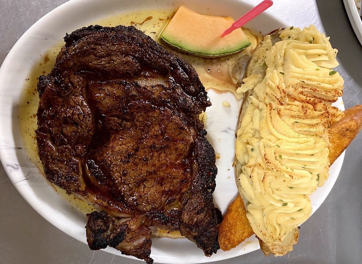 A plate from cheap steak restaurants featuring a grilled steak, a twice-baked potato topped with piped mashed potatoes and paprika, a slice of bread, and a wedge of cantaloupe with a toothpick.