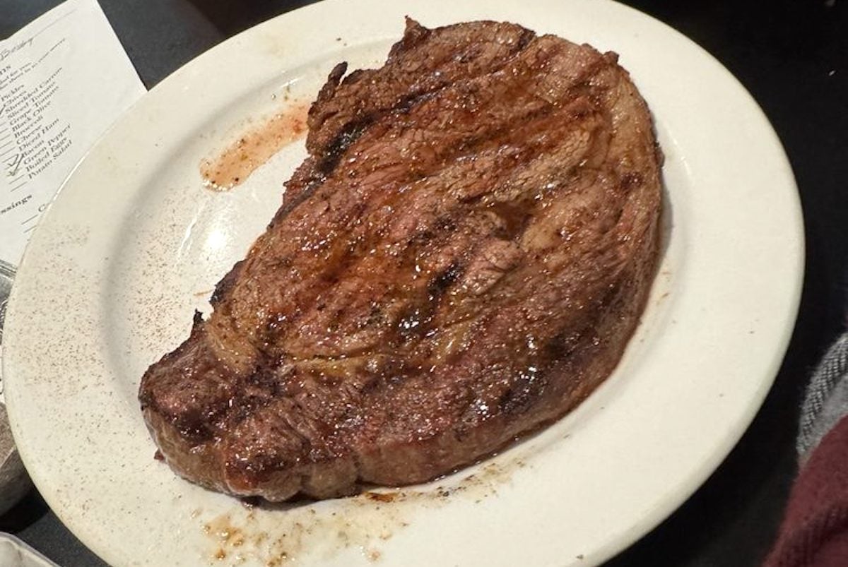 A cooked, grilled steak sits on a white plate, glistening with juices. The steak has visible grill marks and a browned, seared surface—just what you'd expect at many cheap steak restaurants. A menu and a piece of clothing are in the background.