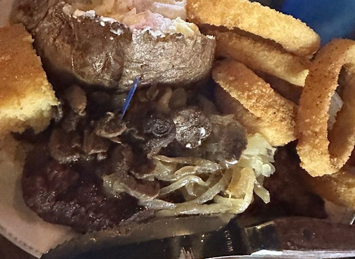 A plate of food from one of the best cheap steak restaurants featuring a baked potato, golden onion rings, sautéed onions, a cooked steak, and a piece of bread with a knife at the bottom edge of the plate.