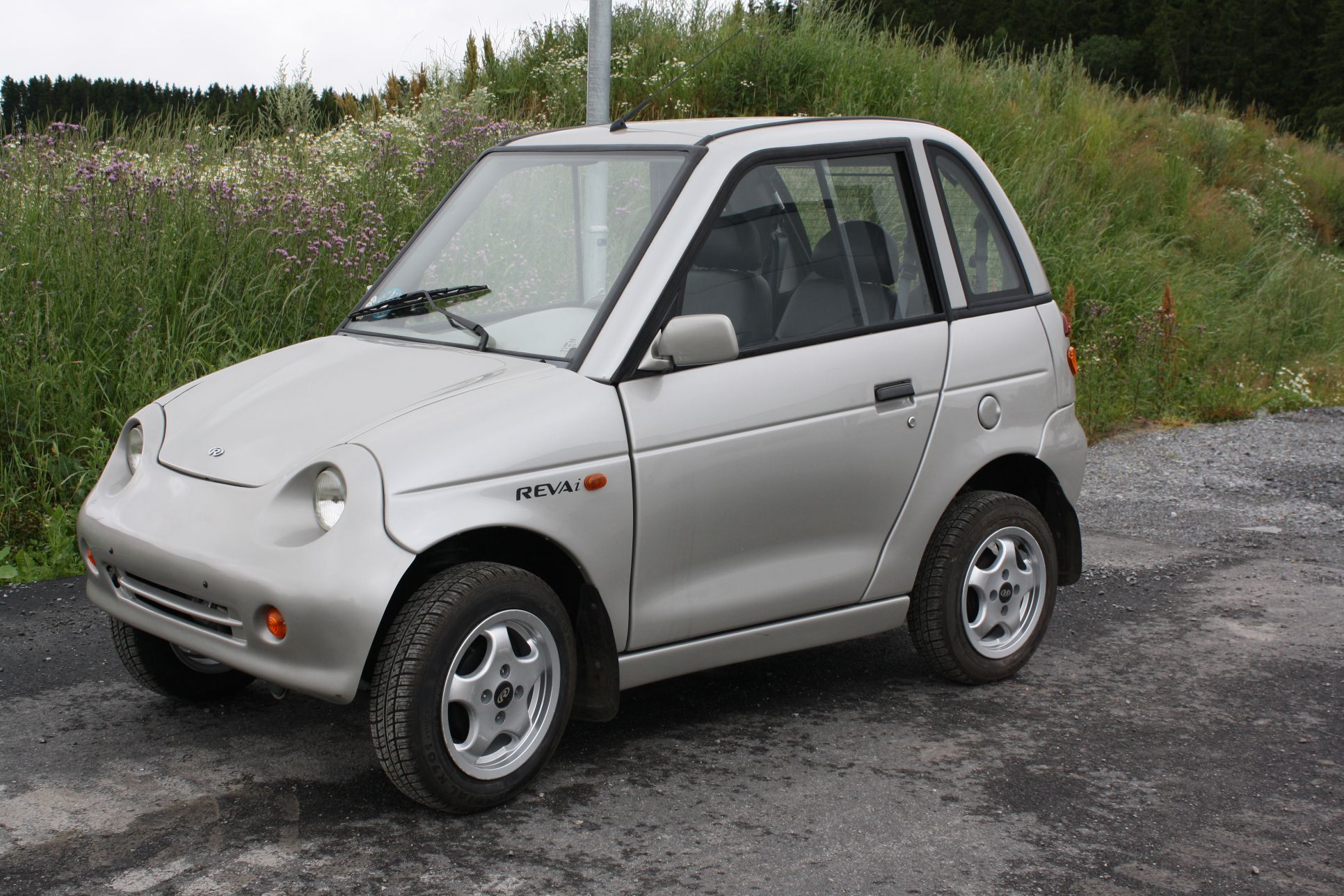 Silver Reva i electric car parked on road with field of wildflowers in background