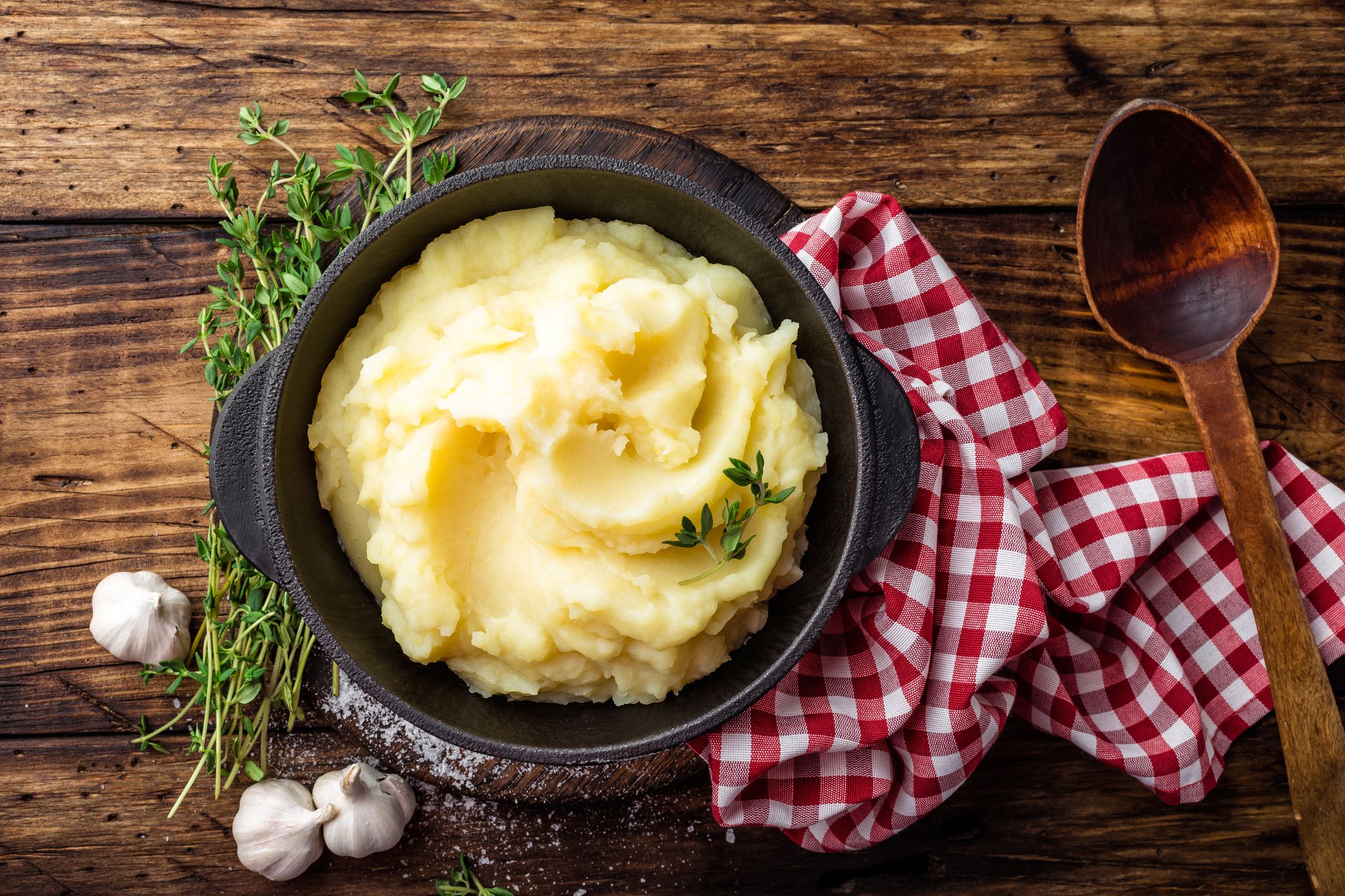 Roasted garlic mashed potatoes in a cast iron pot with a red checkered napkin and a wooden spoon on a rustic wooden table
