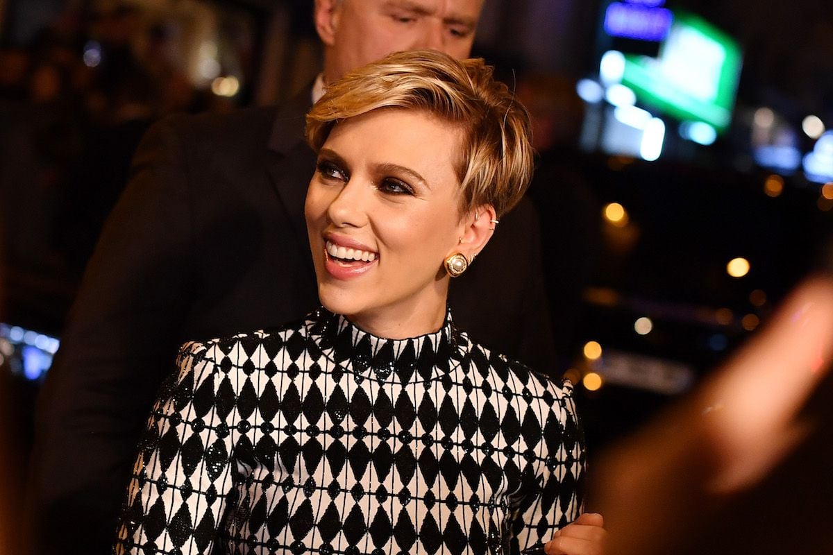 A woman with short blonde hair wearing a black and white patterned dress and gold earrings smiles while standing in a crowd at a nighttime event.