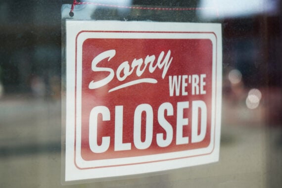 A red and white sign hanging in a window reads "Sorry, we're closed," reflecting how many stores are closed on Thanksgiving 2025. The sign is attached with string, and glass reflections are visible in the background.