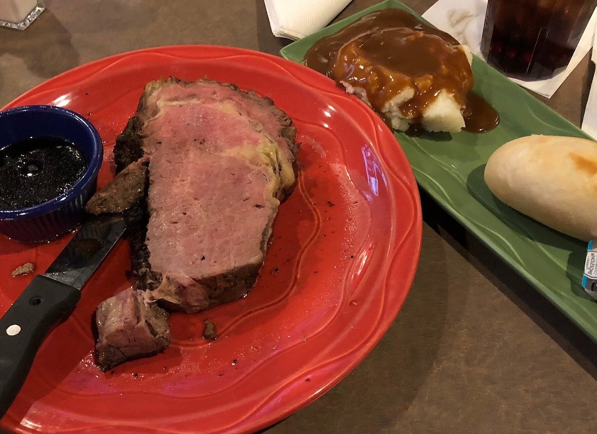 A slice of prime rib with au jus in a small cup on a red plate, accompanied by a knife—classic fare at cheap steak restaurants. Next to it, mashed potatoes with brown gravy and a warm dinner roll with butter on a green tray.