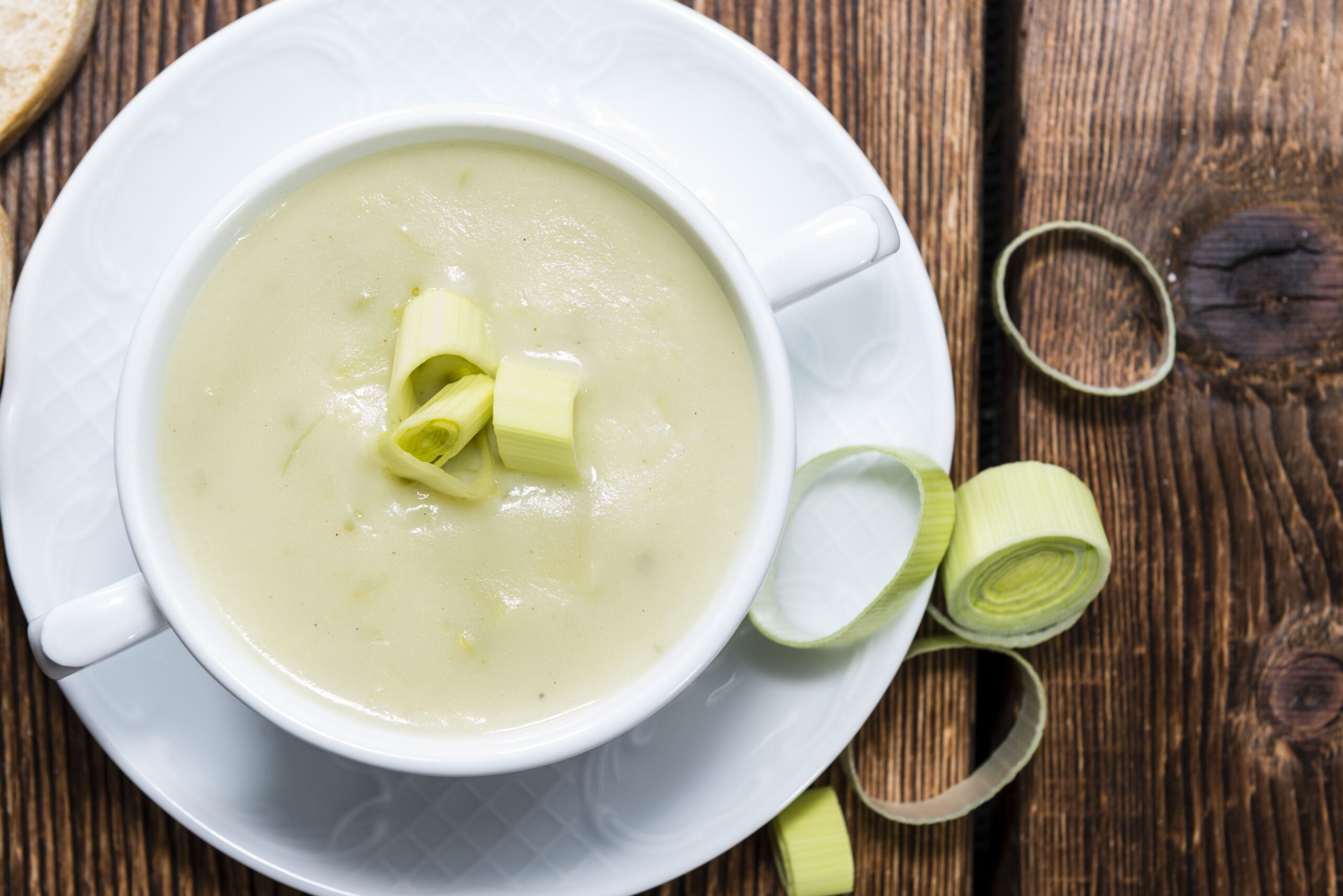 Top view of thick and creamy potato leek soup in a white porcelain bowl on a matching white porcelain plate with leeks on a rustic wooden table