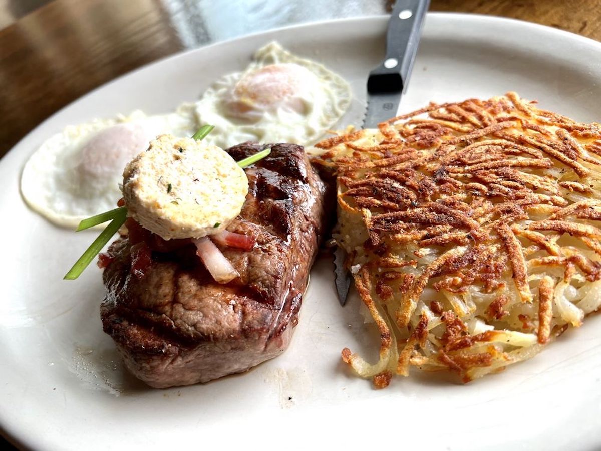 A plate with a grilled steak from one of the best cheap steak restaurants, topped with herb butter and garnish, two fried eggs, and a large serving of crispy hash browns. A steak knife rests on the plate.
