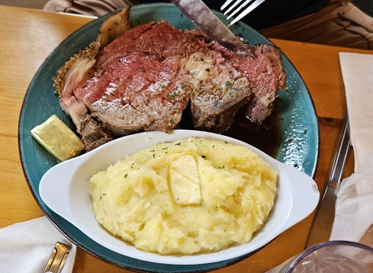 A plate with a thick slice of rare prime rib being cut, served alongside creamy mashed potatoes topped with butter, evokes the hearty comfort found at the best cheap steak restaurants, all set on a rustic wooden table.