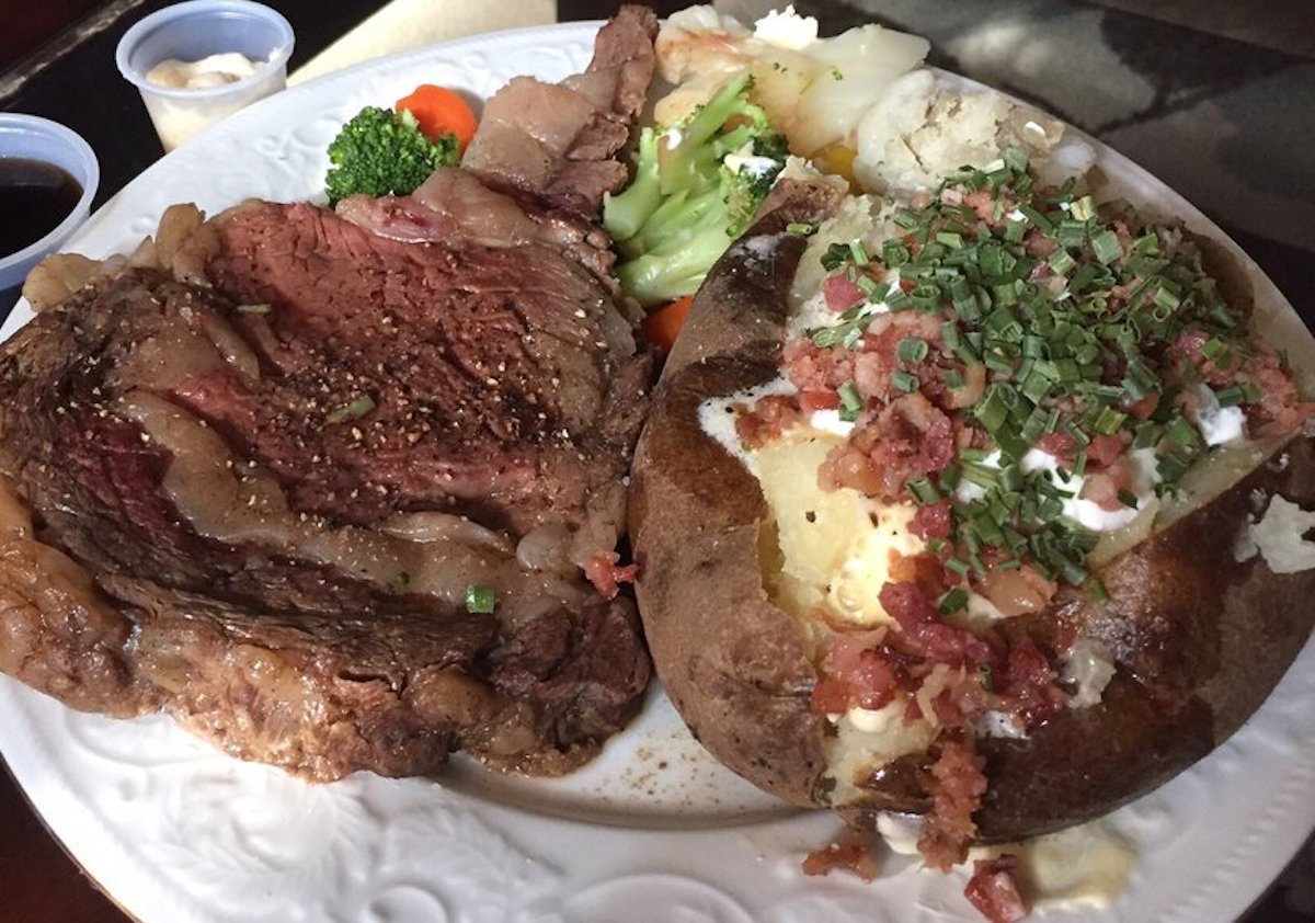 A dinner plate with a large slice of prime rib, like you'd find at cheap steak restaurants, alongside a baked potato topped with sour cream, bacon bits, chives, and a side of steamed broccoli and carrots.