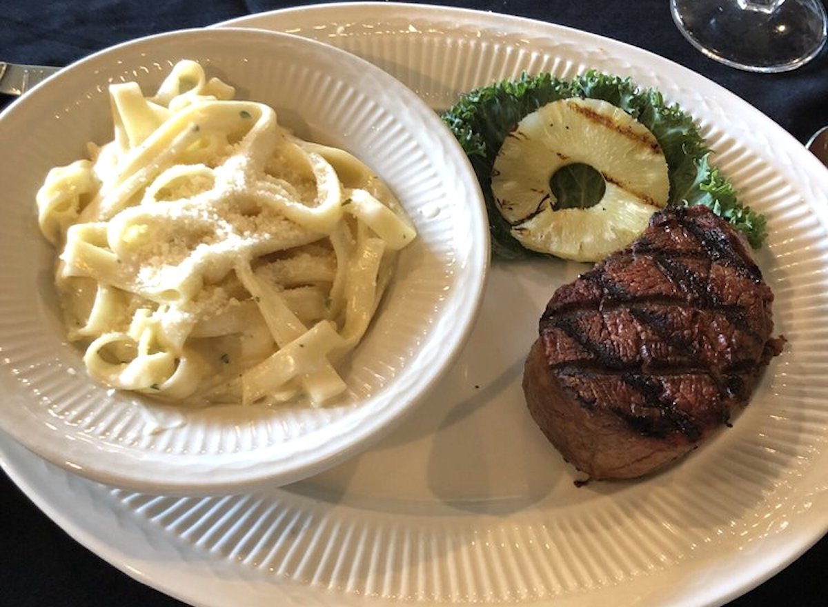 A plate featuring fettuccine Alfredo in a bowl, a grilled steak with char marks from one of the best cheap steak restaurants, a slice of grilled pineapple, and a piece of leafy lettuce on the side.