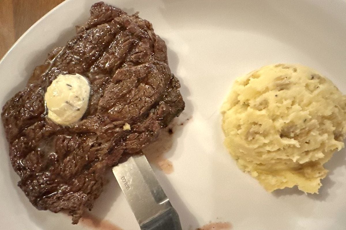 A cooked steak with a pat of herb butter on top sits on a plate next to mashed potatoes flecked with herbs—a classic dish you might find at cheap steak restaurants. A knife rests under the steak.