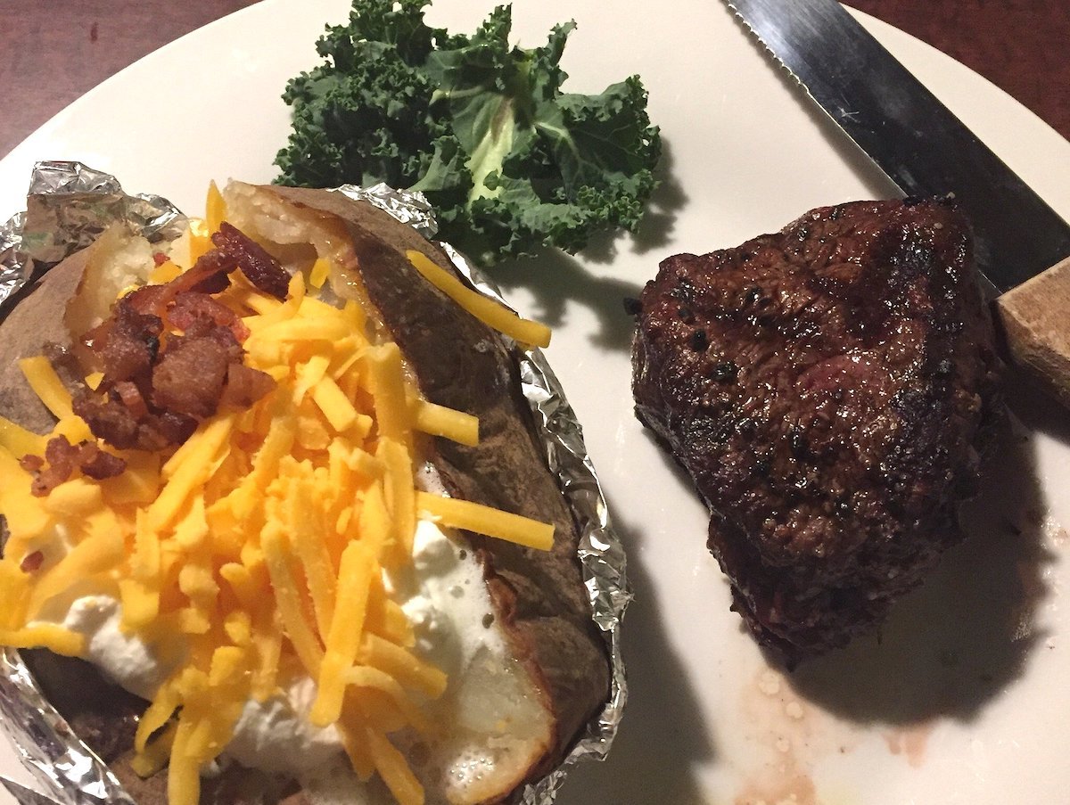A plate with a baked potato topped with sour cream, shredded cheddar cheese, and bacon bits sits next to a grilled steak—classic comfort food you’ll find at many cheap steak restaurants—served with kale and a steak knife.
