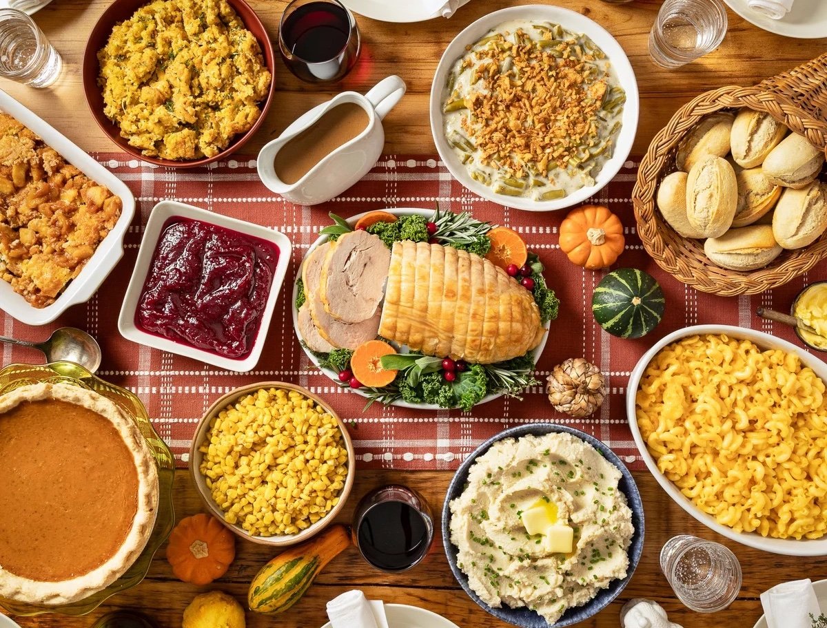 A Thanksgiving meal spread on a wooden table with turkey, mashed potatoes, Costco Thanksgiving sides like macaroni and cheese and green bean casserole, cornbread, cranberry sauce, bread rolls, pie, drinks, and small gourds as garnish.