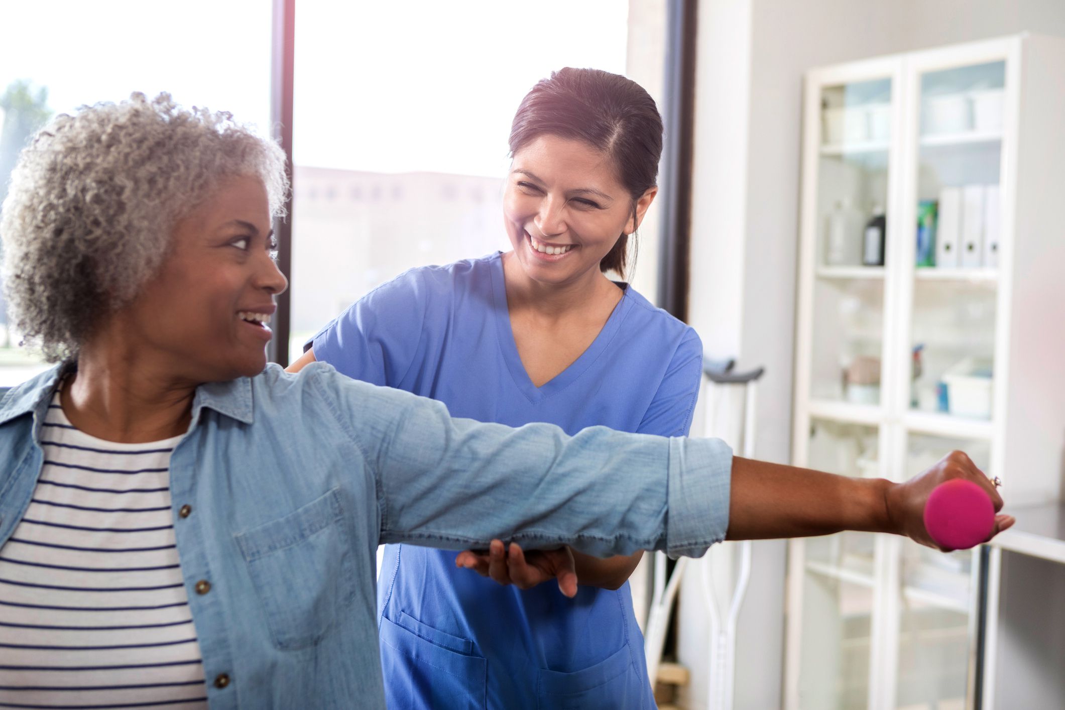 A smiling senior woman sits in a physical therapy office. She reaches out to exercise her arm with a hand weight as her cheerful mid adult female physical therapist stands behind her and guides her arm.