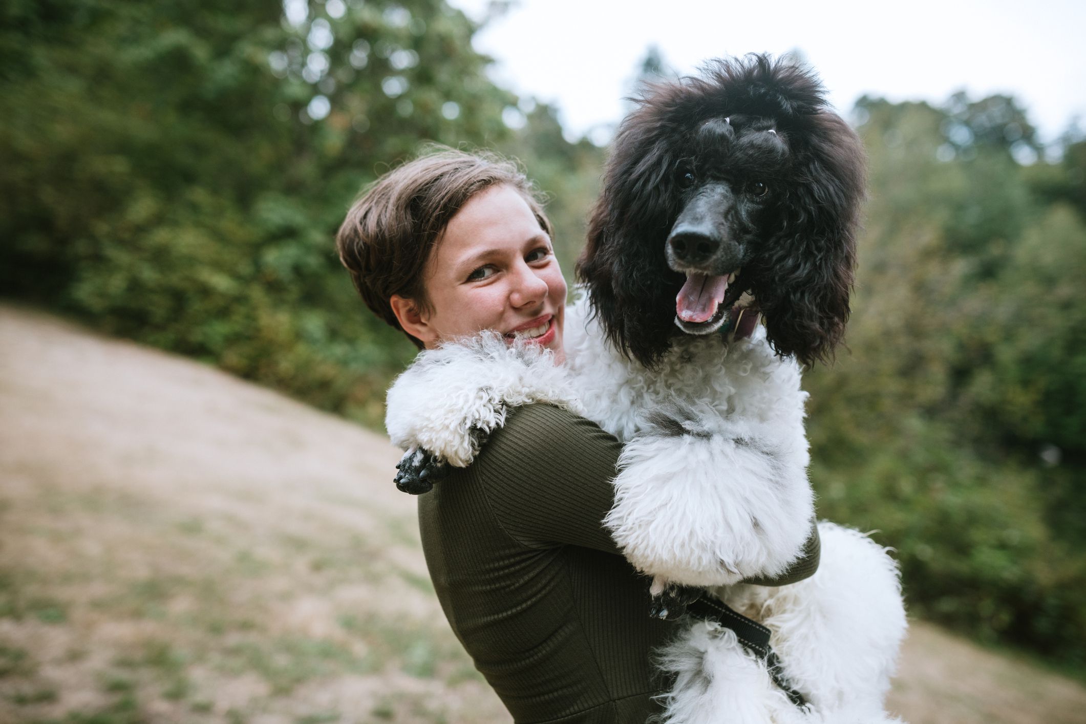 A Caucasian young adult woman with her standard poodle at a park, enjoying the company of her beloved pet. She holds the large dog in a loving embrace. Shot in Washington State, USA.
