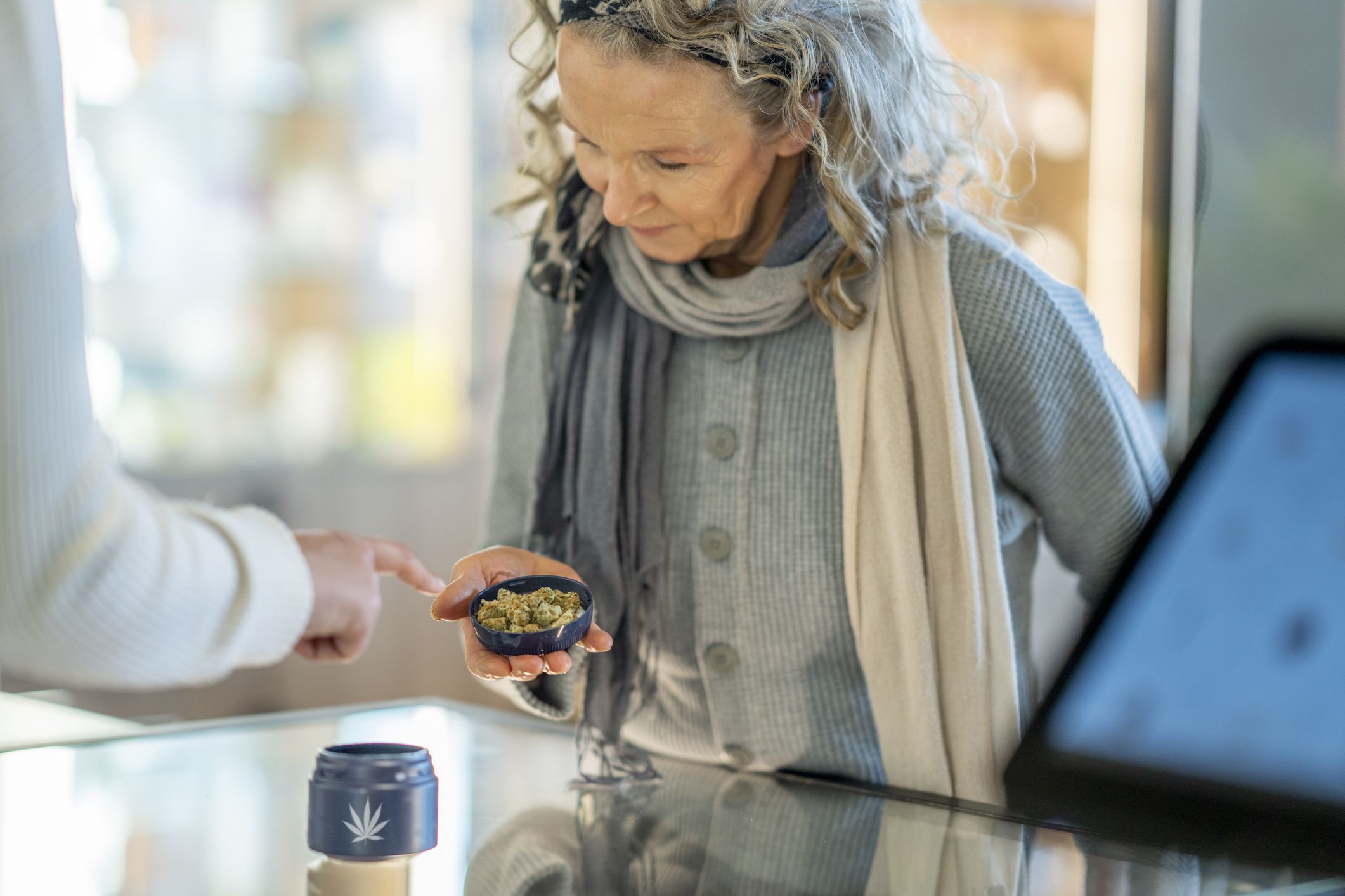 A mature woman stands at the counter of a legal cannabis retailer as she holds out a lid of buds. She is dressed casually and looking down to inspect the product as the associate tries to sell it to her.