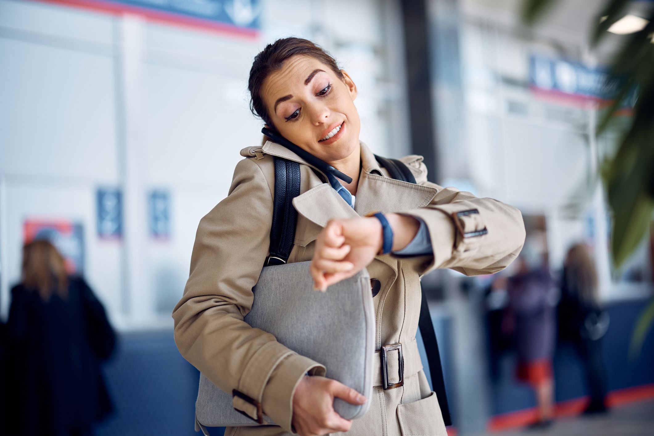 Young businesswoman talking on cell phone and checking the time on her wristwatch while waiting for her transportation at departure area.