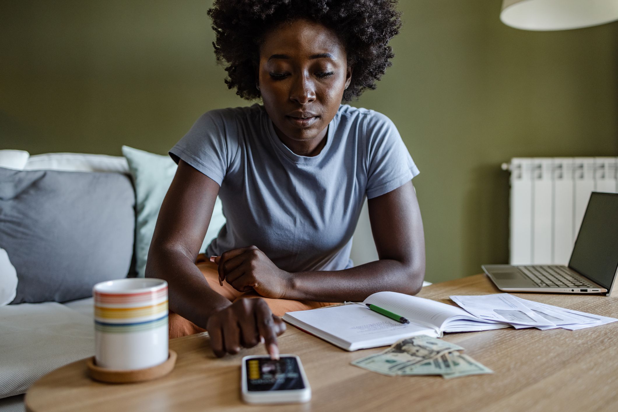 Young African American woman at home doing home budgeting. Woman is sitting on the couch and calculating expenses on her phone.